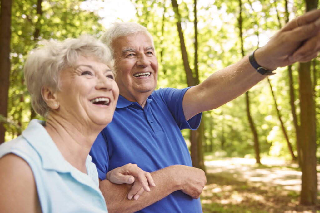 deux séniors souriants, un homme, une femme, au milieu d'une forêt en pleine journée, des arbres tout au tour.
l'homme indique quelque chose au loin à la femme