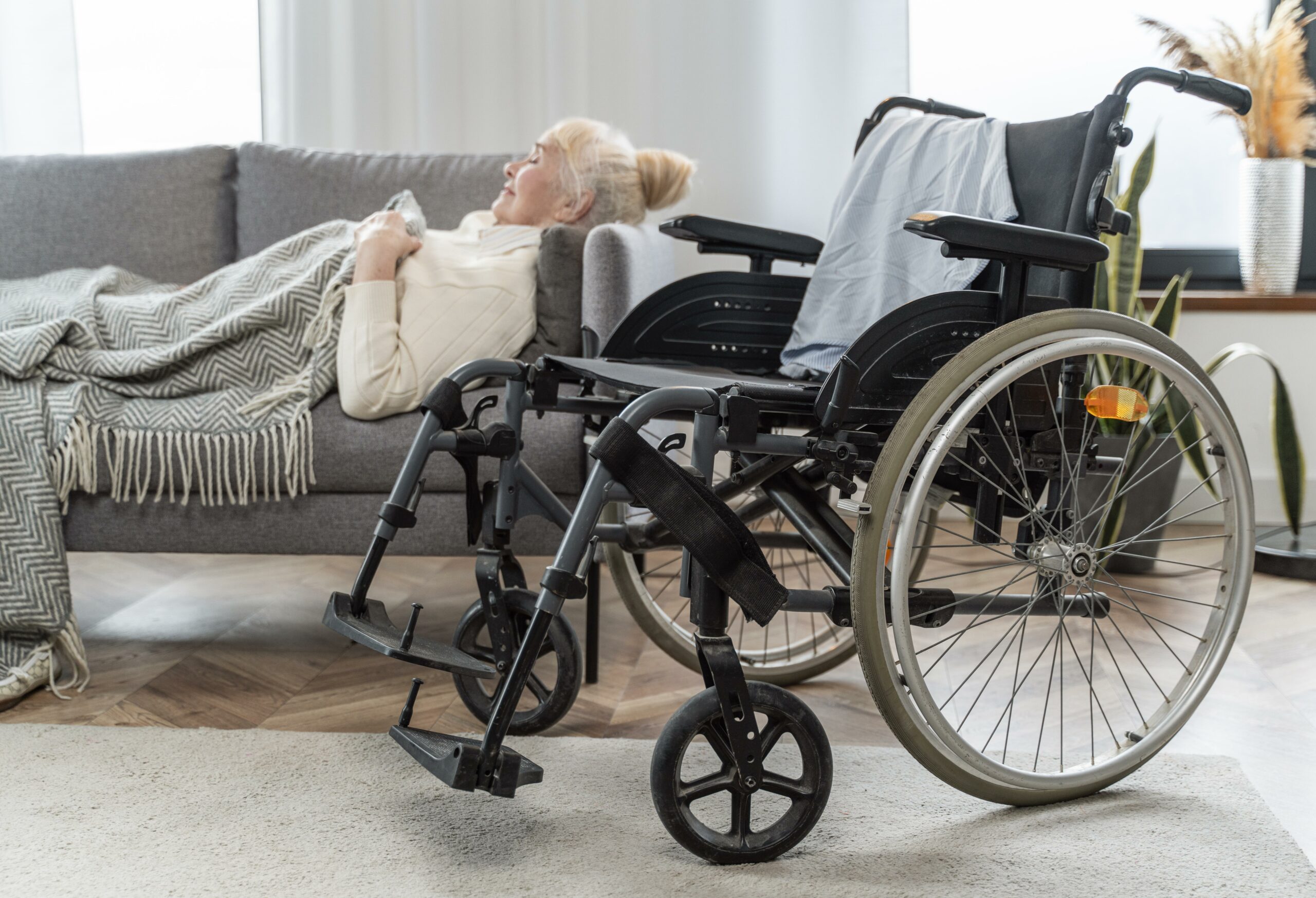 une femme sénior allongée dans un canapée. Elle est recouverte d'un plaid et semble dormir. Au premier plan, il y a un fauteuil roulant positionné de 3/4